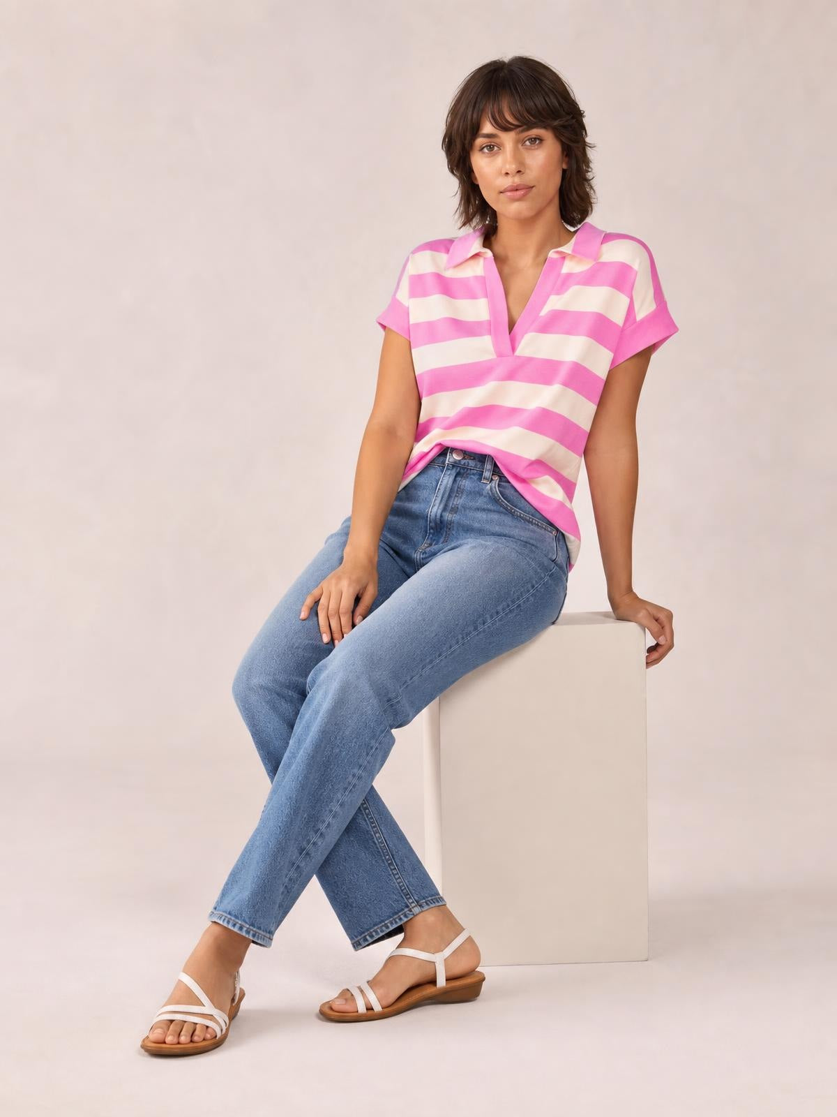 Model seated in a relaxed pose wearing a pink and white striped notched cap sleeve blouse with a loose, straight silhouette and rolled sleeve cuffs.