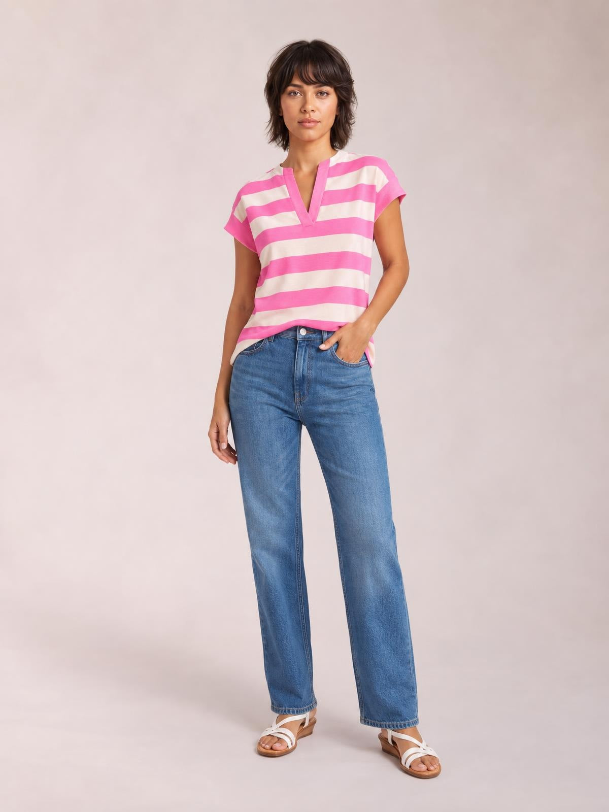 Model wears a pink and ivory striped blouse with a notched V-neck, cap sleeves, and a relaxed straight silhouette, posing with one hand in her jeans pocket.