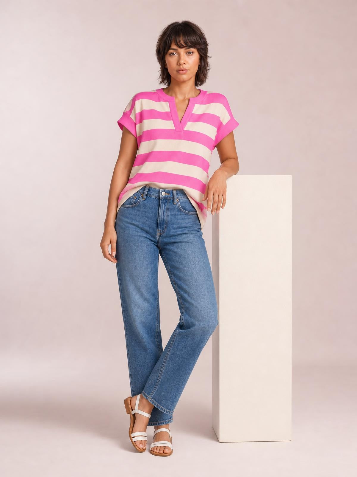 Model wearing a pink and cream striped notched cap sleeve blouse with a relaxed silhouette, standing with one hand resting on a pedestal.