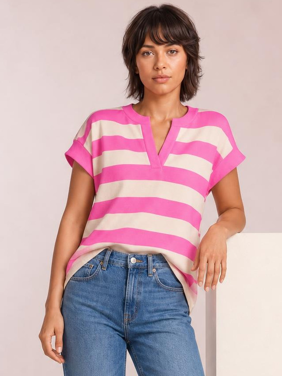 A model poses in a relaxed fit striped blouse with bold pink and ivory bands, a notched neckline, cap sleeves, and a straight silhouette.