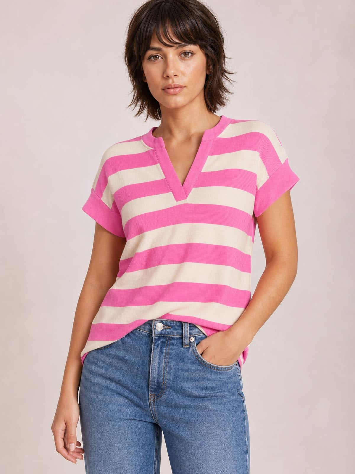Model wearing a pink and cream striped notched cap sleeve blouse with a relaxed silhouette, standing with one hand in her jeans pocket.