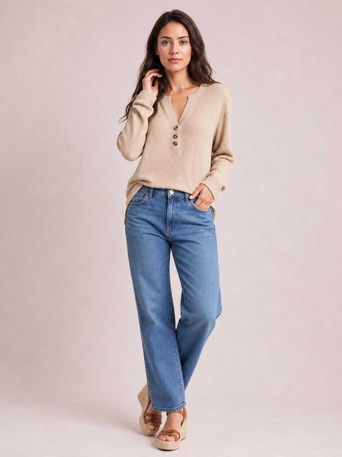 A model wears a beige long-sleeve t-shirt with a notched neckline, ribbed texture, three-button placket, and relaxed silhouette, posing with one hand at her hair.