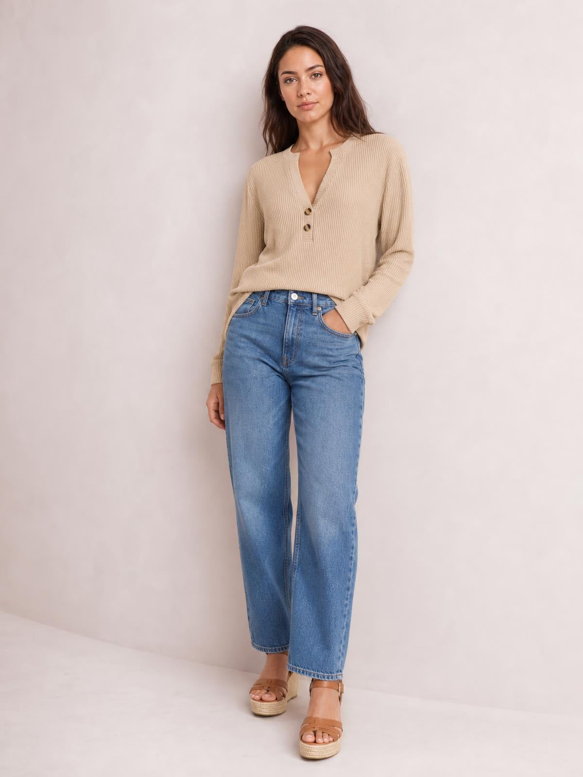Model wears a beige long sleeve top with a notched V-neck, ribbed construction, button accents, and a relaxed silhouette with one hand in her jeans pocket.