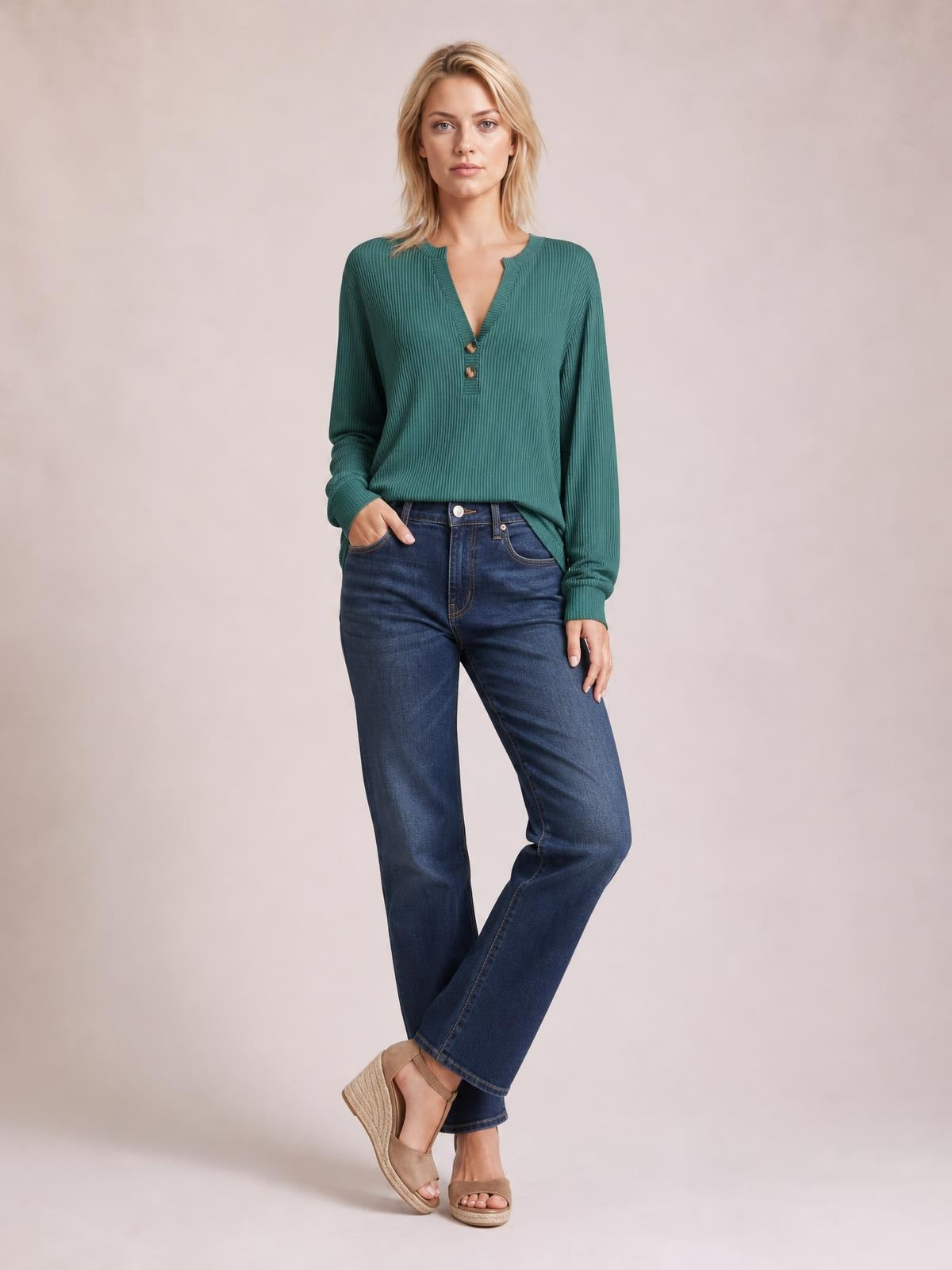 A model wears a green striped long sleeve T-shirt with a notched neckline, button accents, ribbed texture, and a relaxed silhouette while standing with crossed ankles.