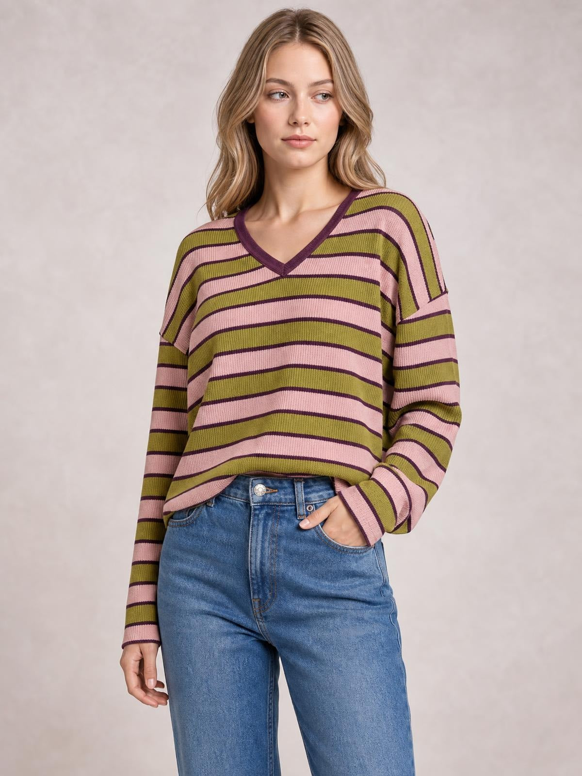 Model wears a relaxed striped notched long sleeve T-shirt in olive green, pink, and purple with a V-neckline and gently oversized silhouette.
