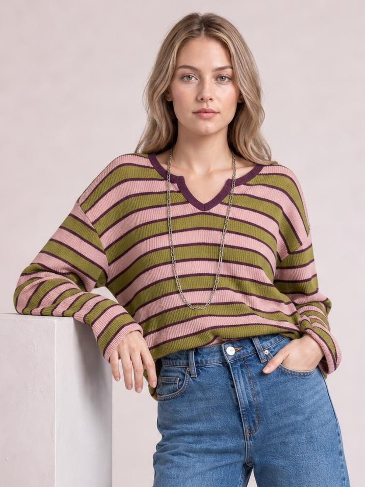 A model wears a pink and olive striped notched long sleeve T-shirt with a relaxed silhouette, resting one hand on a ledge and the other in her jeans pocket.