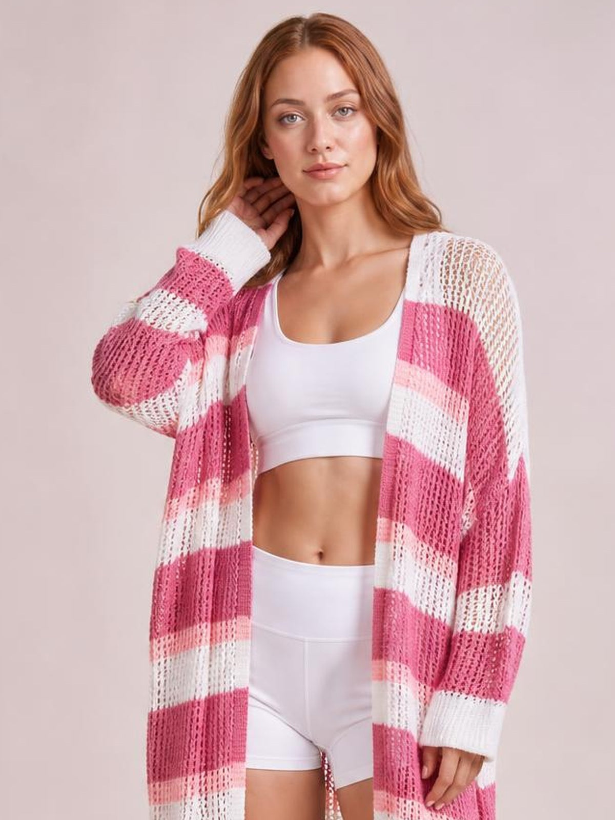 A model poses in a sheer pink-and-white striped open-front duster cover-up with long sleeves, an easy longline silhouette, and a hand lifted to her hair.