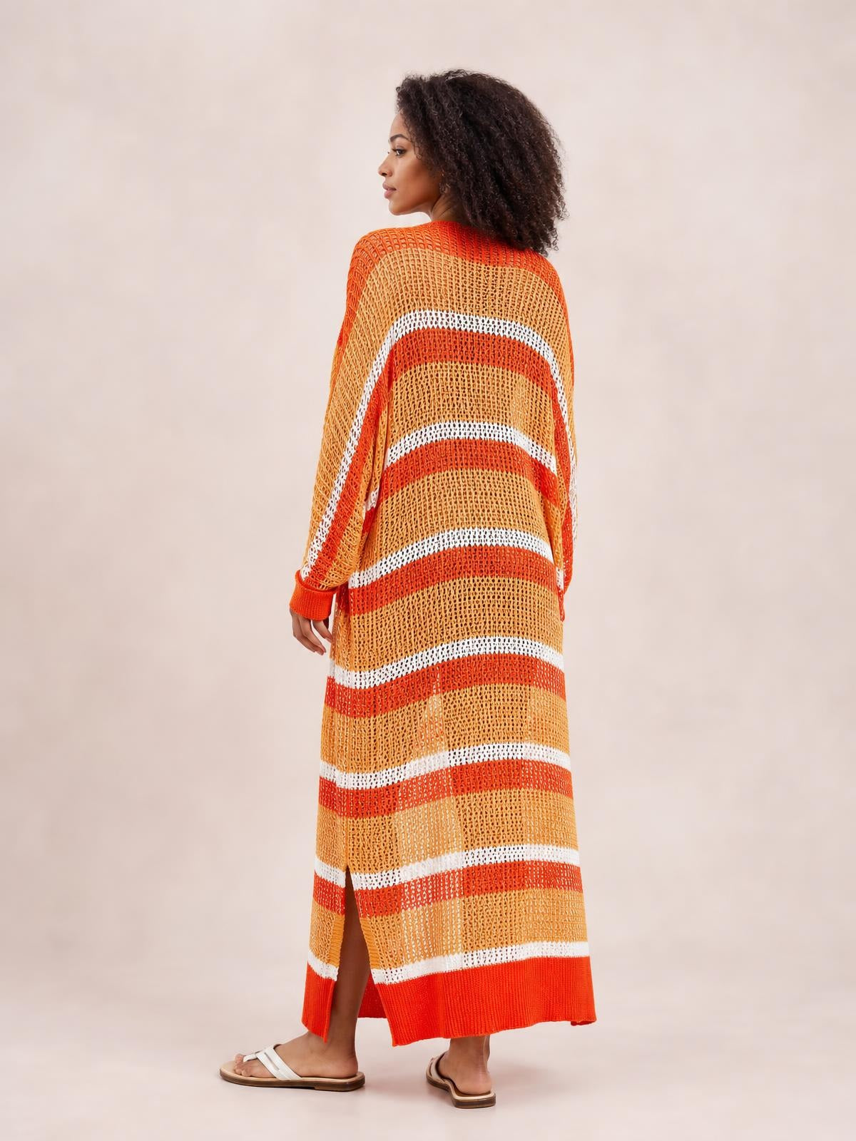 A model stands turned away in a sheer orange, tan, and white striped open-front duster cover up with long sleeves, a relaxed long silhouette, and side slits.