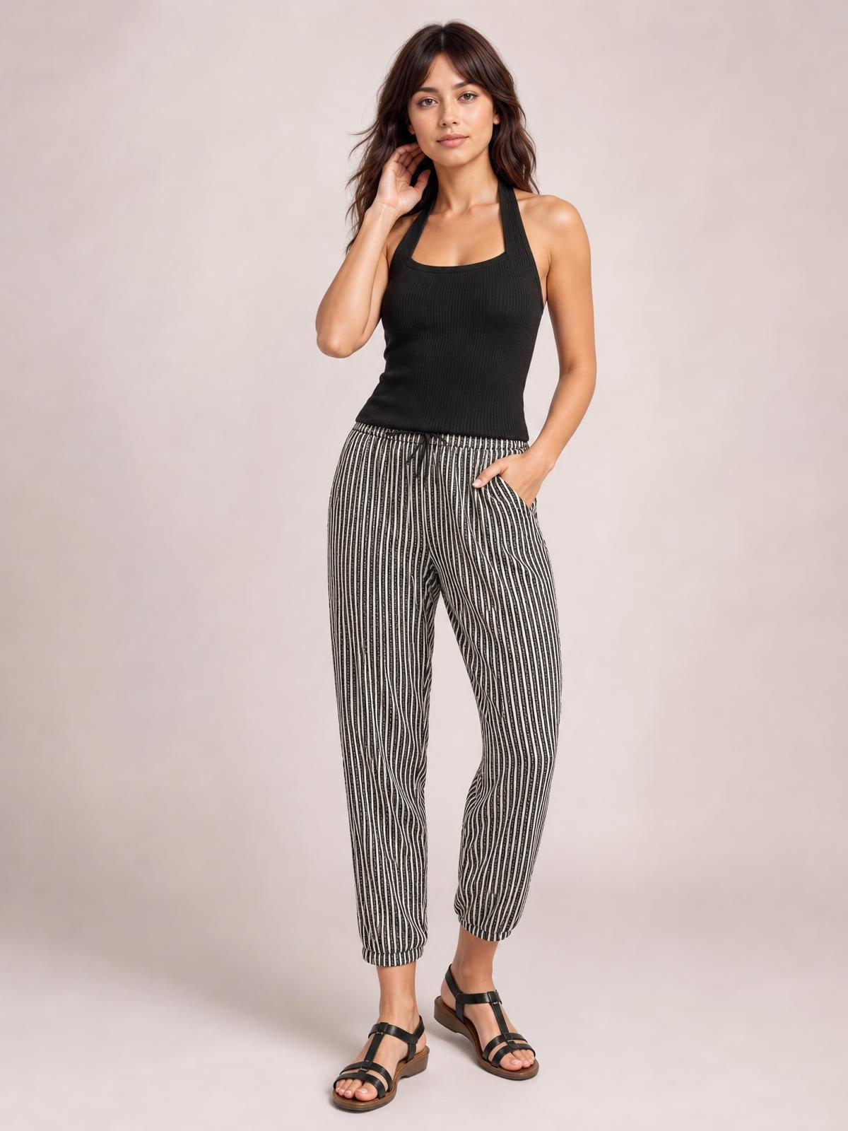 A model poses in black-and-white striped pants with a relaxed tapered silhouette, gathered waist, cuffed hems, and side pockets, one hand tucked inside.
