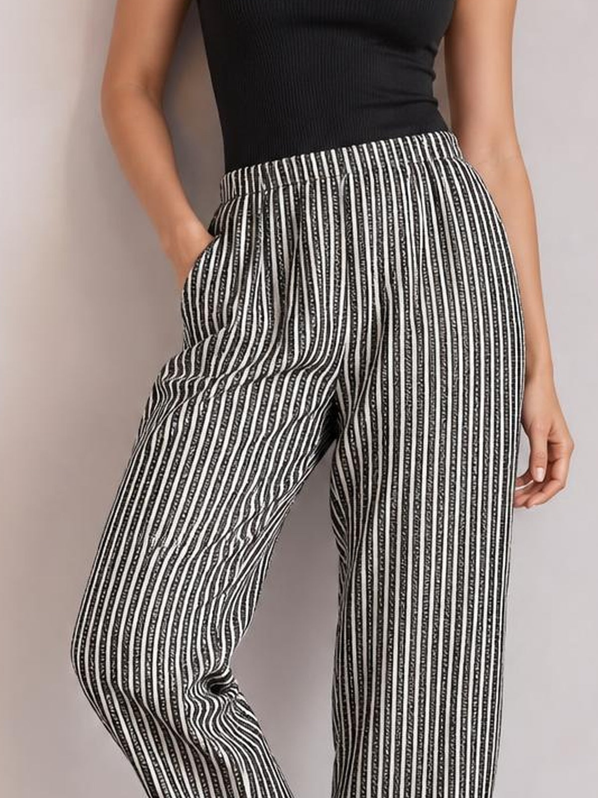 A visible model poses with one hand in the pocket, wearing black-and-white vertically striped pants with an elastic waistband and relaxed straight-leg silhouette.