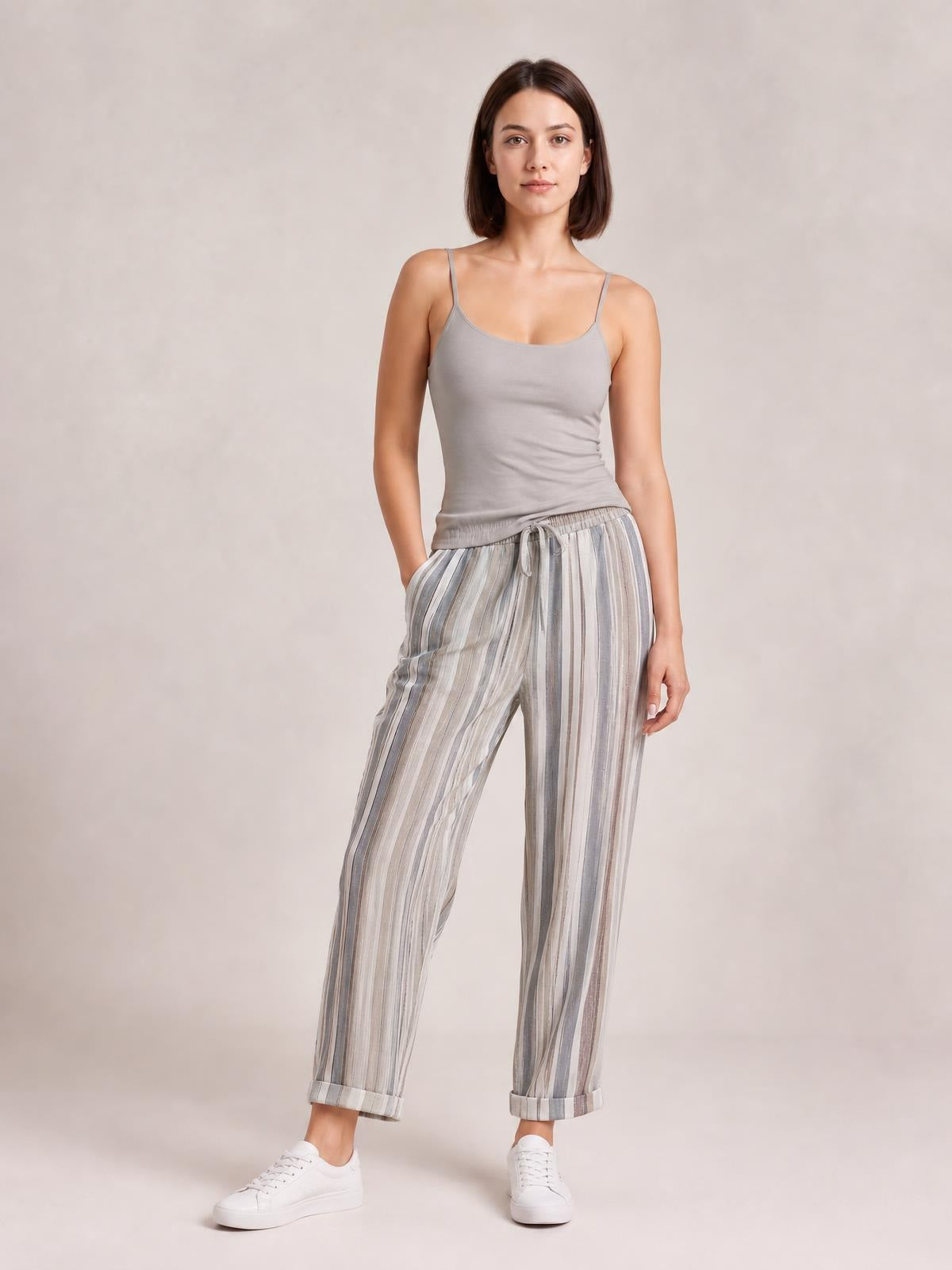 A woman models striped pants with gray and white vertical stripes, a relaxed straight-leg silhouette, drawstring waist, side pockets, and cuffed hems.