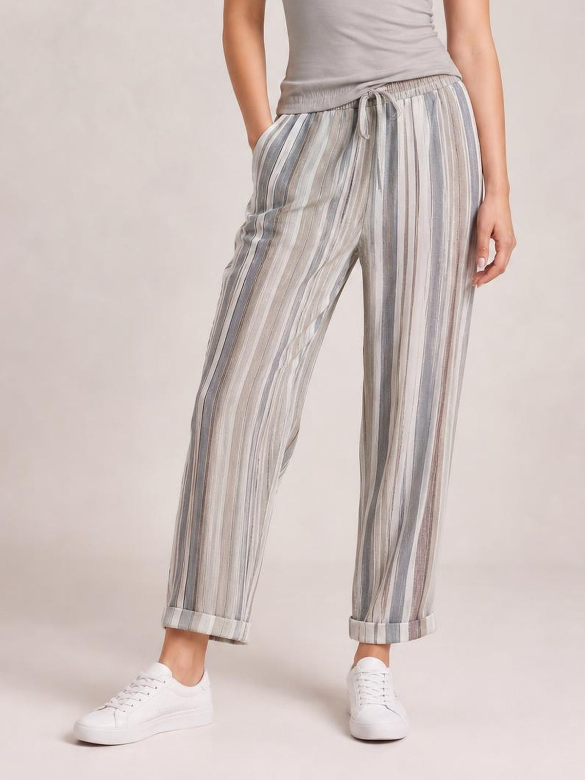 A model wears opaque striped pants with pockets, an elastic drawstring waist, relaxed straight-leg silhouette, and cuffed hems in muted gray and white tones.
