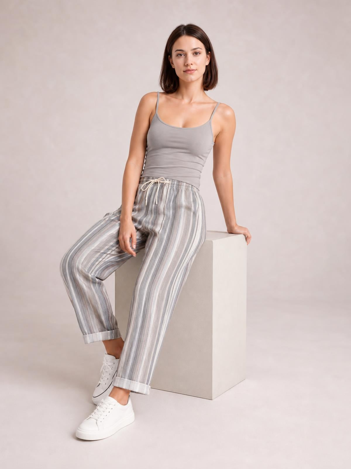 Model seated and leaning against a block wears gray and white vertically striped pants with pockets, a drawstring waist, relaxed tapered legs, and cuffed hems.