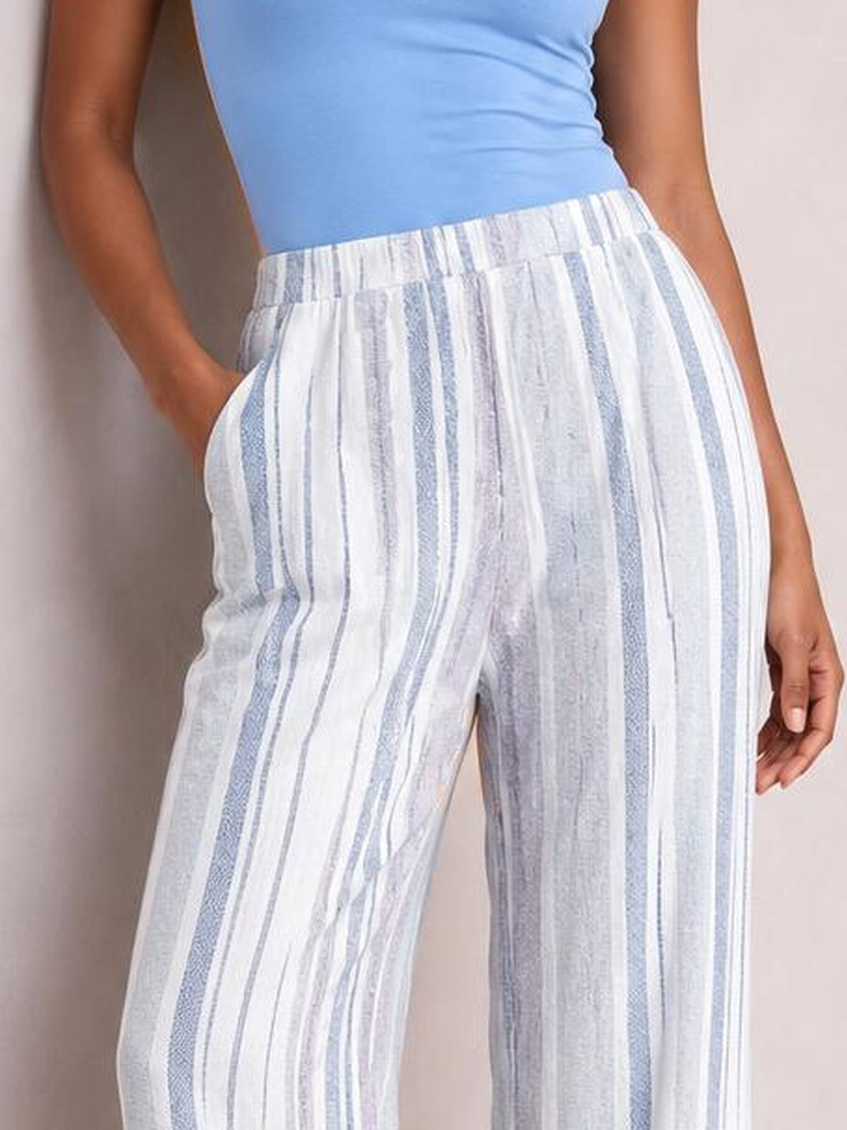 Model wears white and light blue striped pants with a high elastic waistband, relaxed straight-leg silhouette, and visible side pockets, standing with one hand tucked inside.