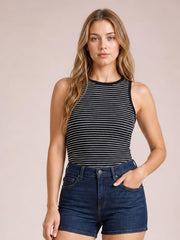 A model wears a fitted black striped ribbed tank with a round neckline and sleeveless silhouette, posing front-facing with one hand in her denim shorts pocket.