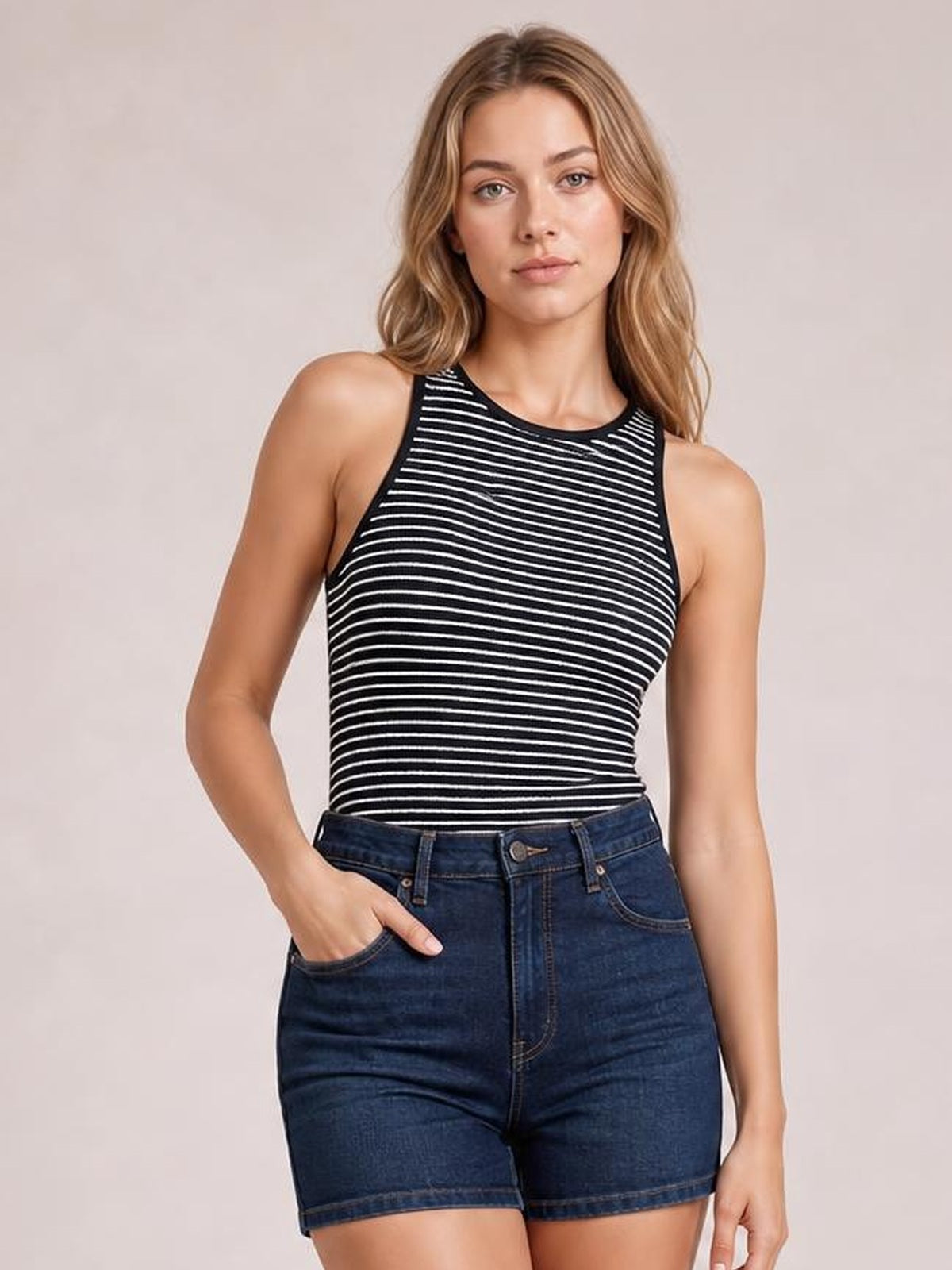 Model wearing a black-and-white striped ribbed round neck tank with a fitted silhouette and wide shoulder straps, posed with one hand in her denim shorts pocket.