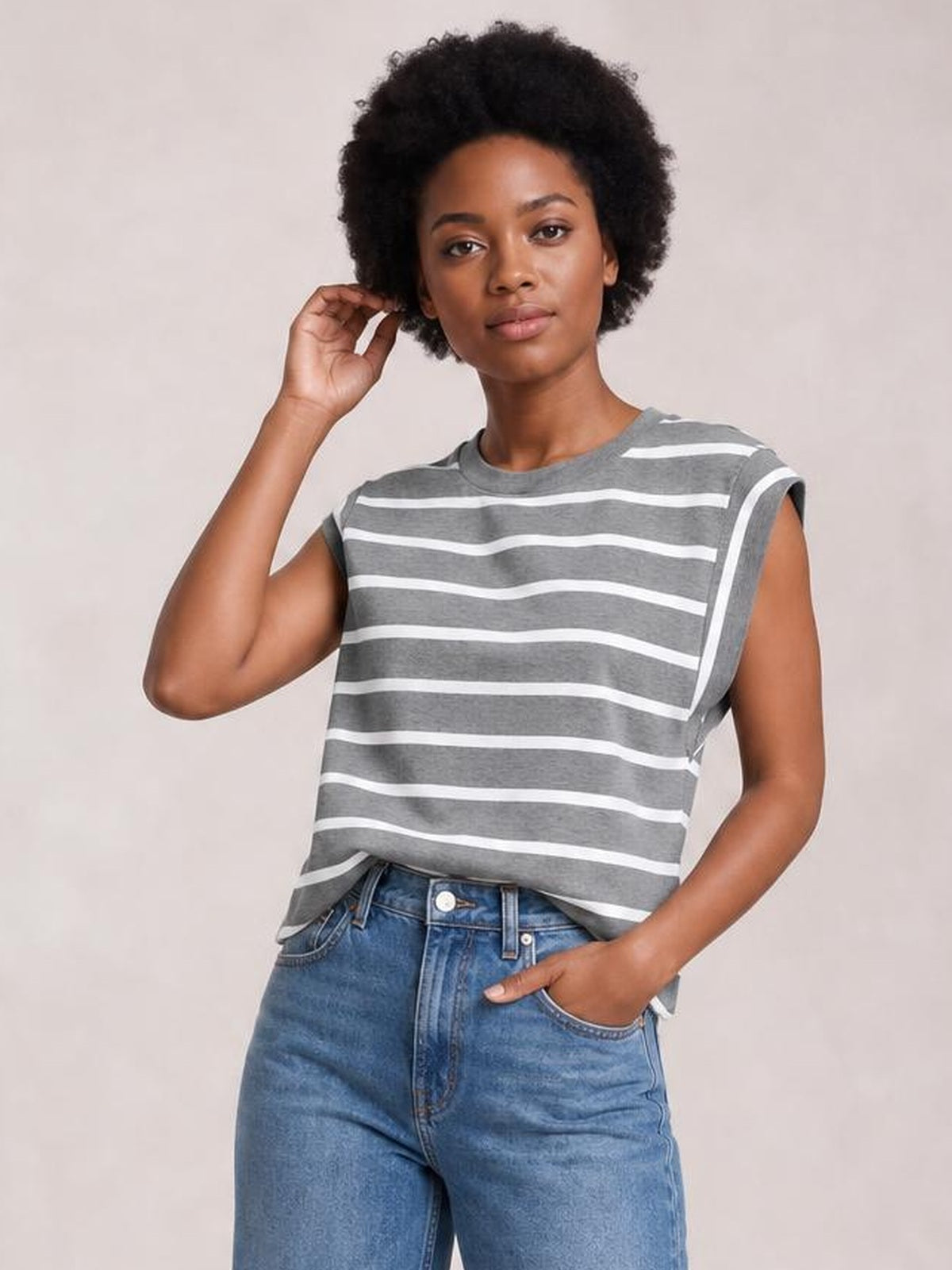 Model poses in a gray and white striped round neck cap sleeve T-shirt with a relaxed silhouette, one hand in her jeans pocket and the other touching her hair.