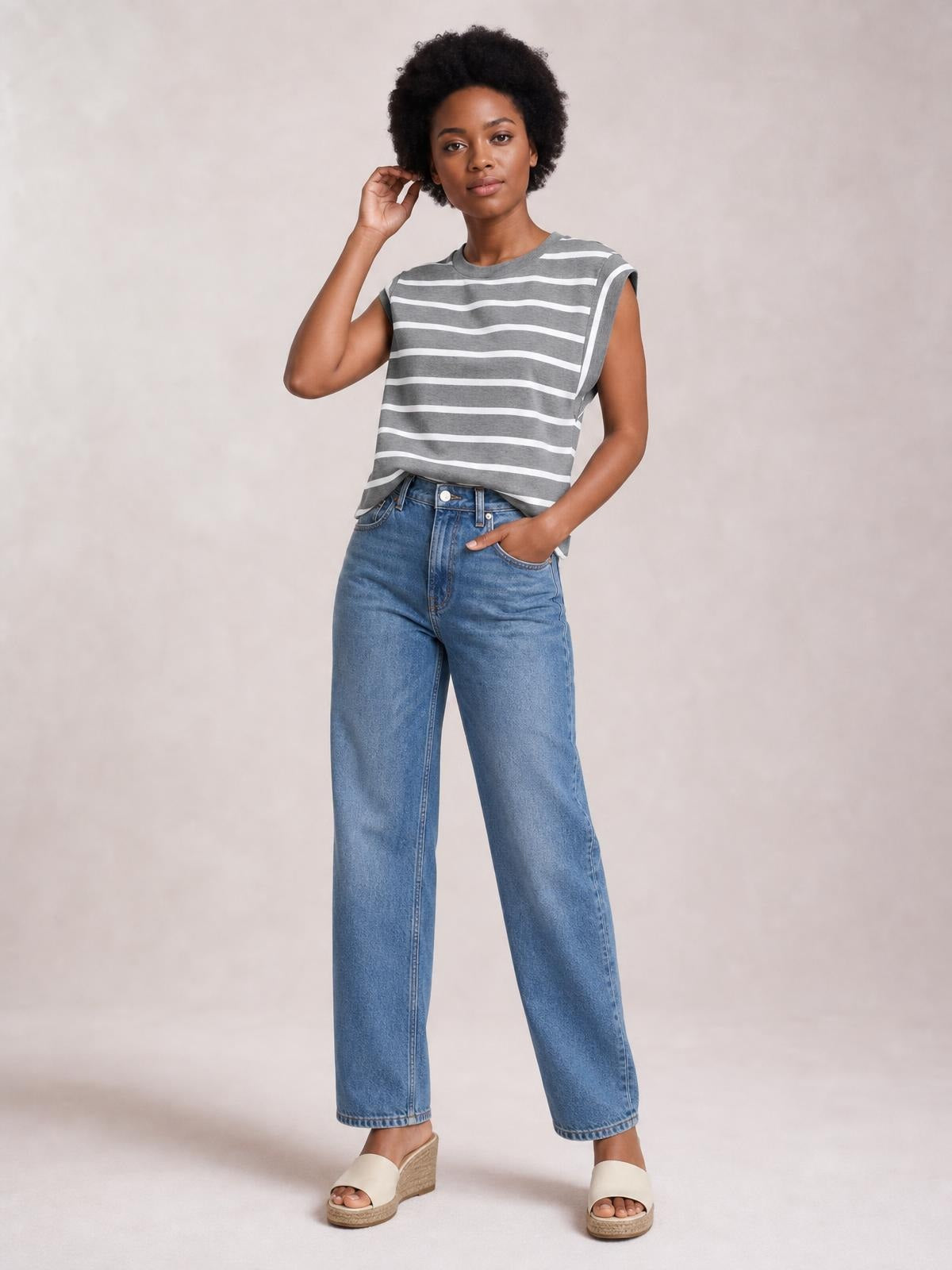 Model wearing a gray and white striped round neck cap sleeve T-shirt with a relaxed silhouette, one hand in her jeans pocket and the other touching her hair.
