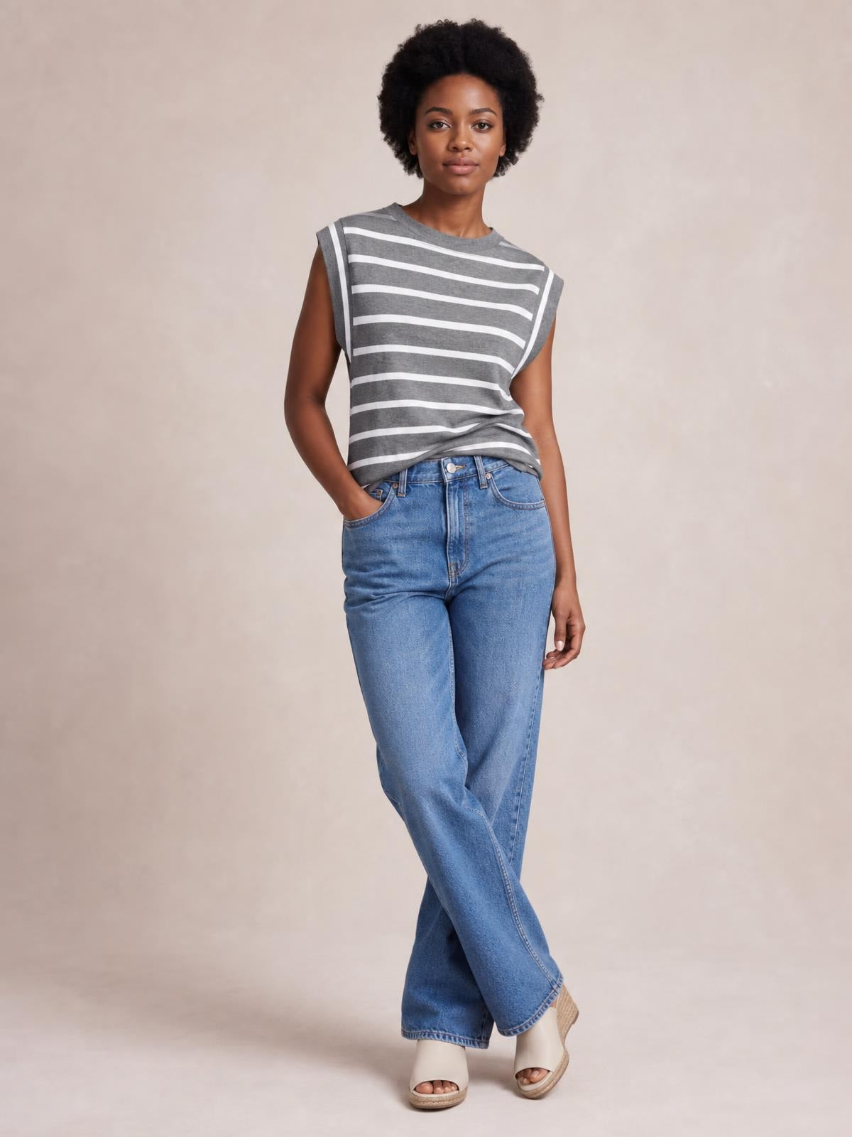 Model poses with one hand in her pocket wearing a gray and white striped round neck cap sleeve T-shirt with a relaxed silhouette and slightly gathered hem.