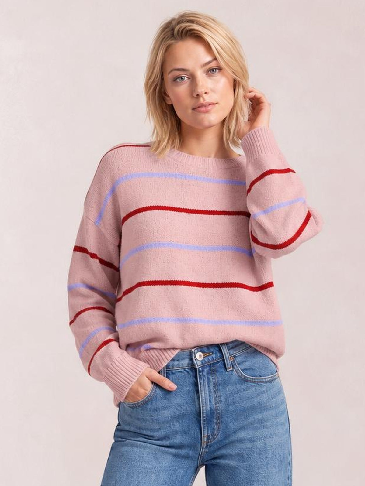 Model wearing a pink striped round neck drop shoulder sweater with red and lavender bands, relaxed silhouette, ribbed cuffs and hem, posing with one hand in a pocket.