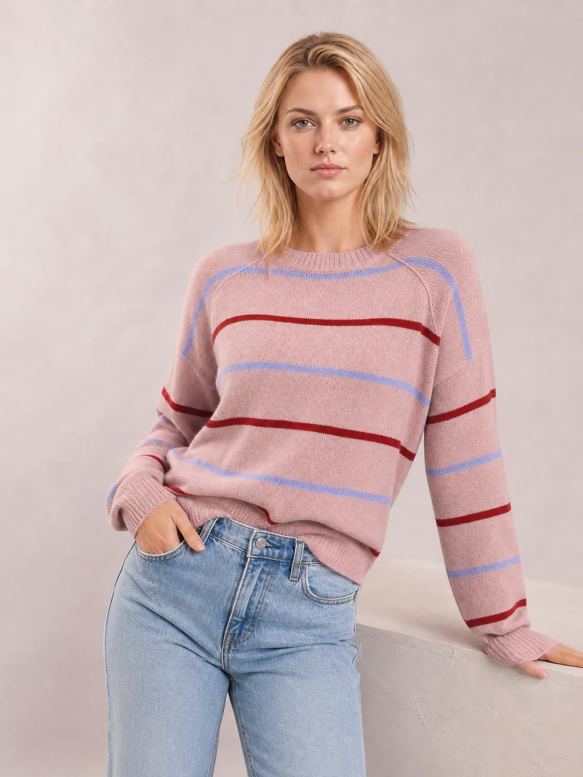 Model wearing a pink striped round neck drop shoulder sweater with blue and burgundy bands, relaxed silhouette, ribbed cuffs and hem, posing with one hand in her jeans pocket.