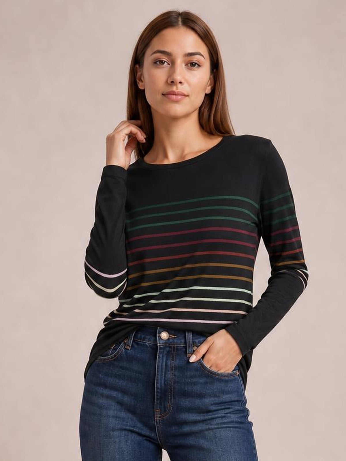 Model wears a black striped round neck long sleeve T-shirt with multicolor horizontal bands, a relaxed silhouette, one hand at her hair and the other in a jean pocket.