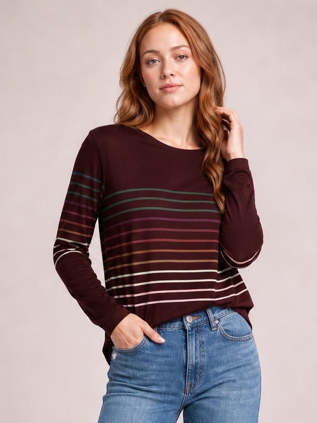 Model poses in a burgundy striped round neck long sleeve T-shirt with a relaxed silhouette, curved hem, and multicolored horizontal stripe detailing.