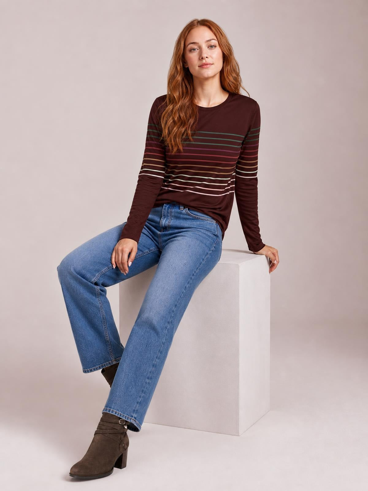 Model seated in a relaxed pose wears a burgundy striped round neck long sleeve T-shirt with a straight silhouette and multicolor horizontal bands.