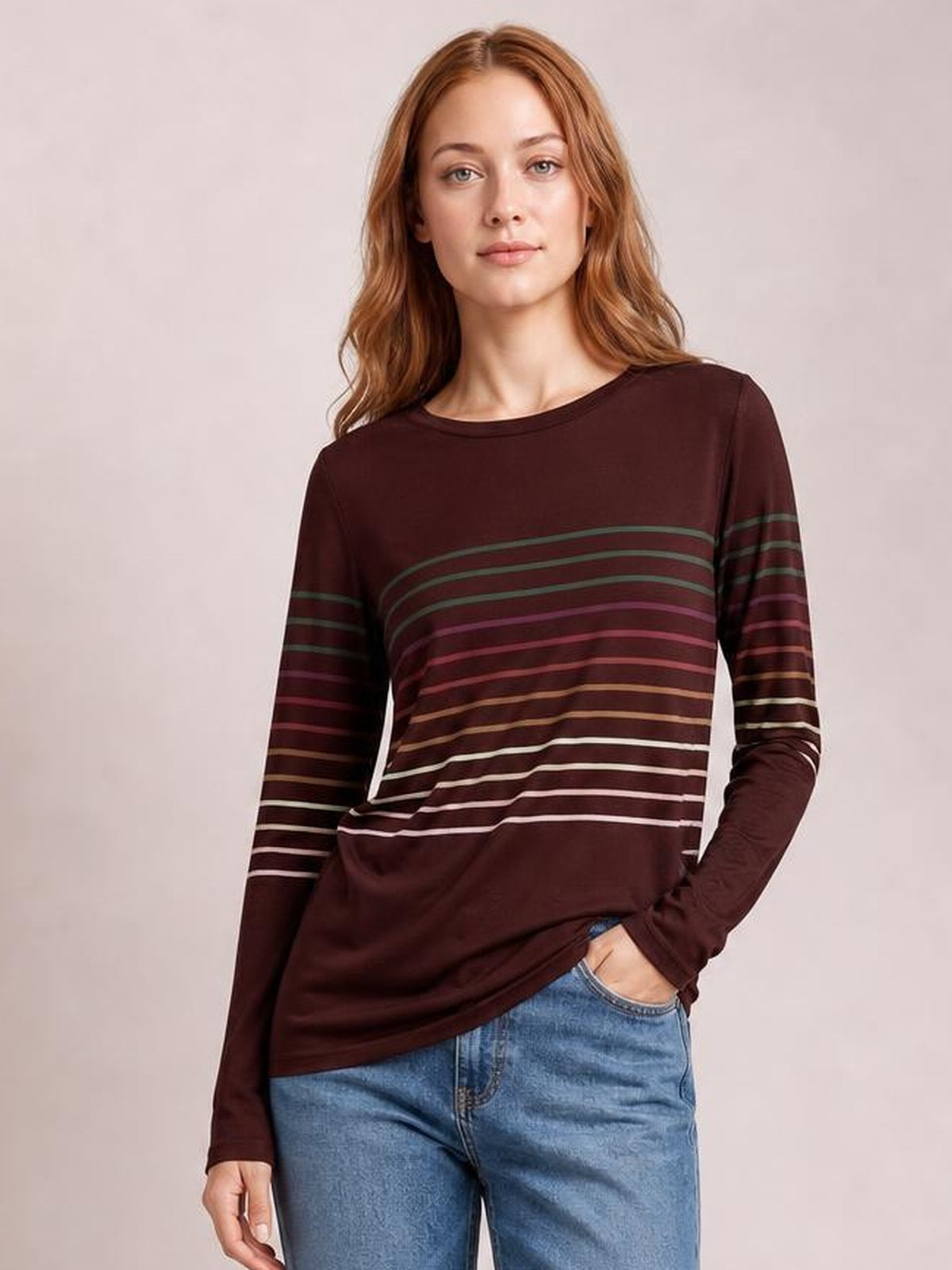 Model wears a burgundy striped round-neck long-sleeve T-shirt with multicolor horizontal bands, a relaxed silhouette, and one hand tucked into her jeans pocket.