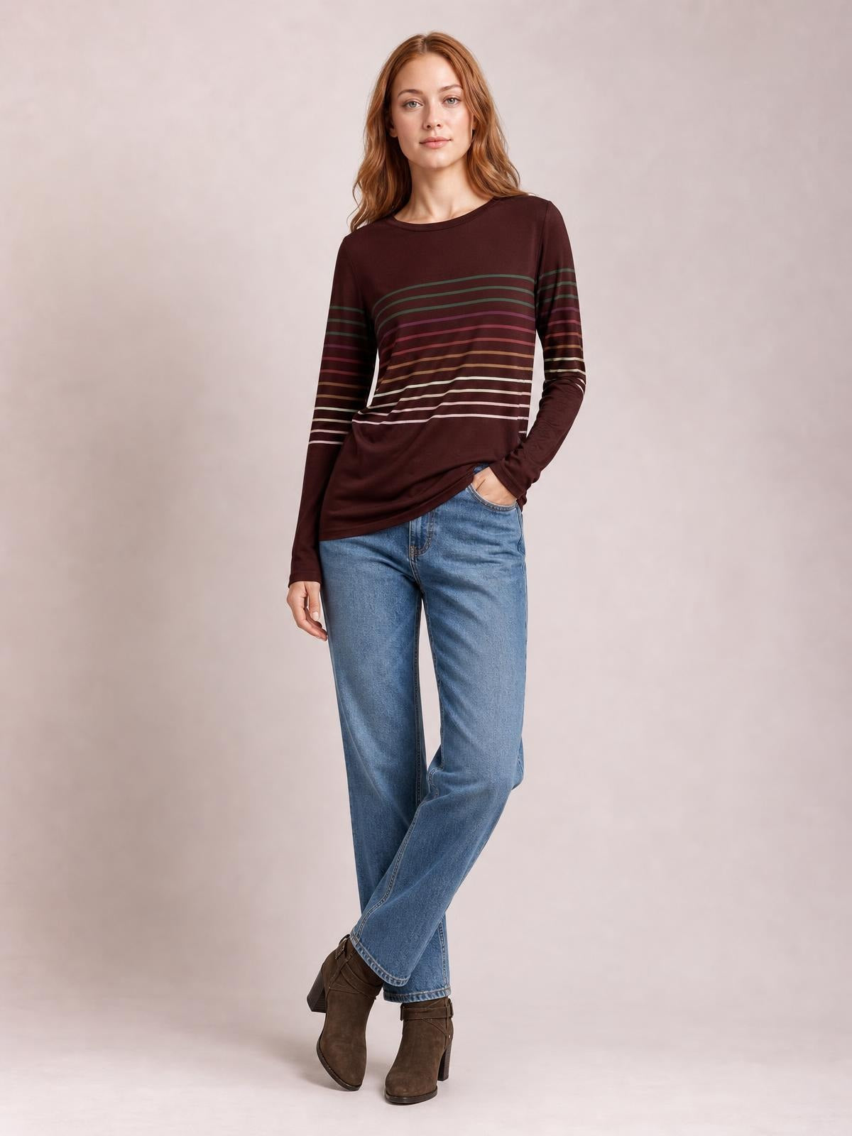 A woman models a burgundy striped round neck long sleeve T-shirt with multicolor horizontal bands, relaxed fit, and a slightly curved hem while standing with crossed legs.