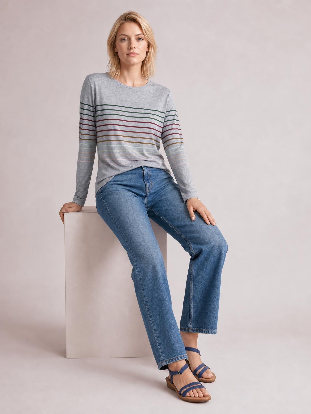 A seated model wears a gray striped round neck long sleeve T-shirt with multicolor horizontal bands and a relaxed, slightly fitted silhouette.