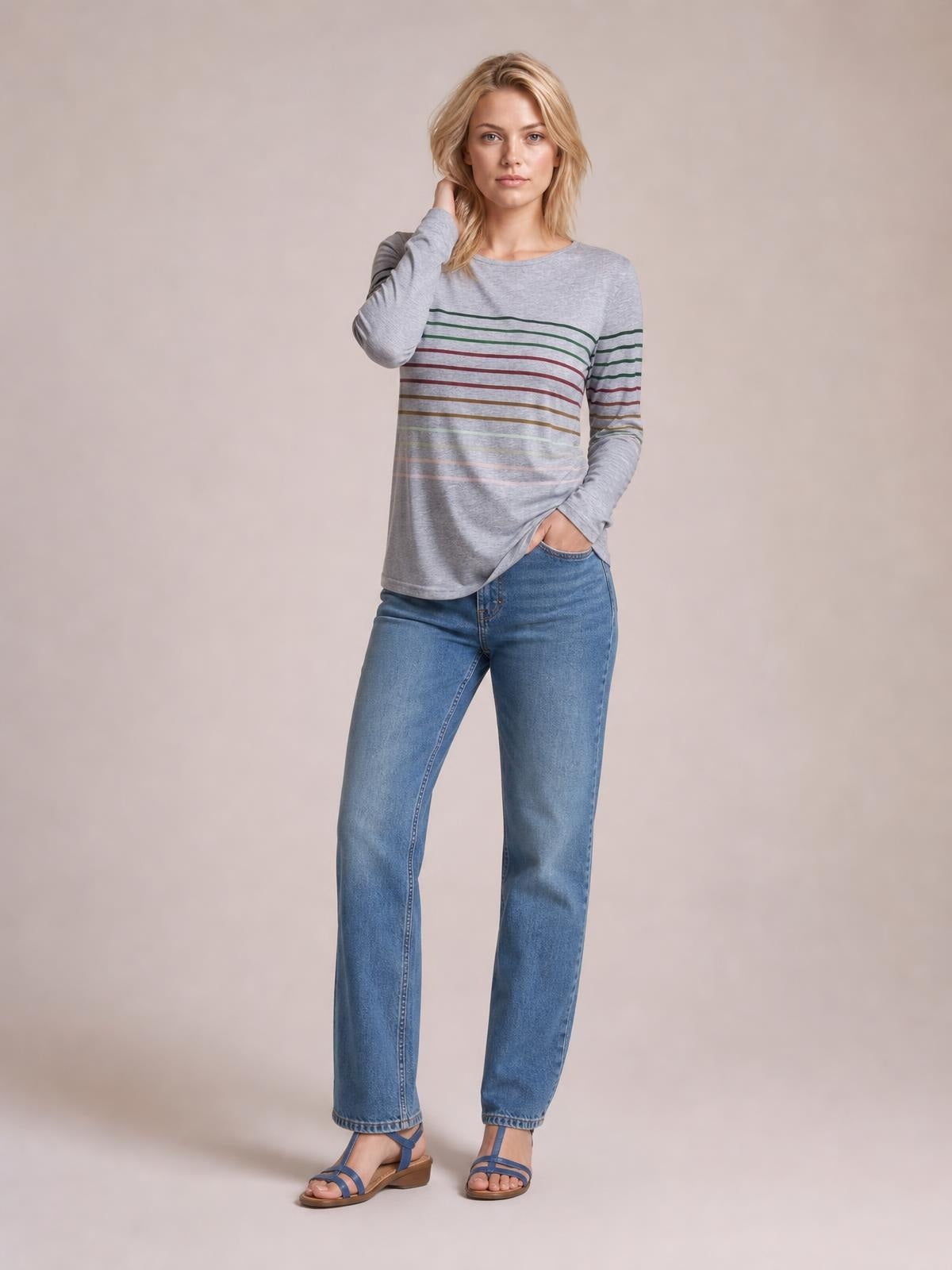 Model wears a gray striped round neck long sleeve T-shirt with multicolored horizontal bands and a relaxed silhouette, posing with one hand in her pocket.