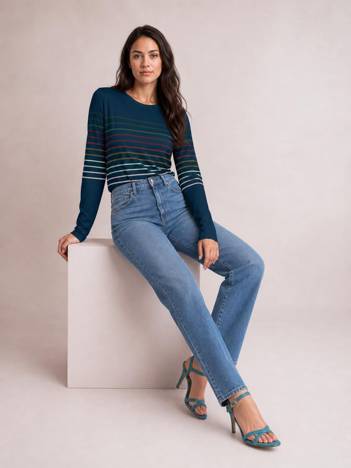 Model seated in a relaxed pose wears a navy striped round neck long sleeve T-shirt with multicolored horizontal bands and a fitted silhouette.