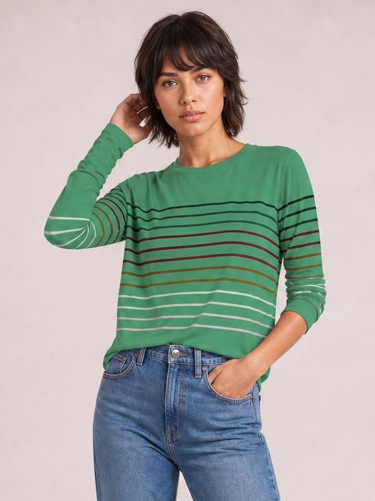 Model wears a green striped round neck long sleeve T-shirt with a relaxed silhouette, posing with one hand in her jeans pocket and the other touching her hair.