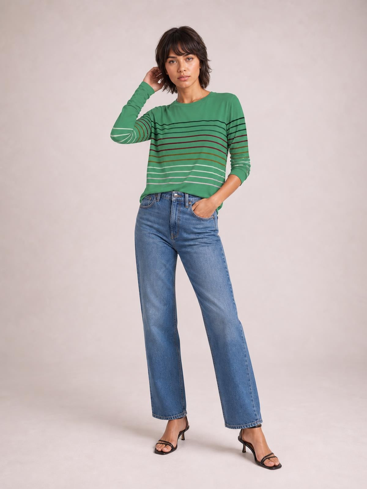 Model wearing a green striped round neck long sleeve T-shirt with a relaxed silhouette, standing with one hand in her jeans pocket and the other touching her hair.