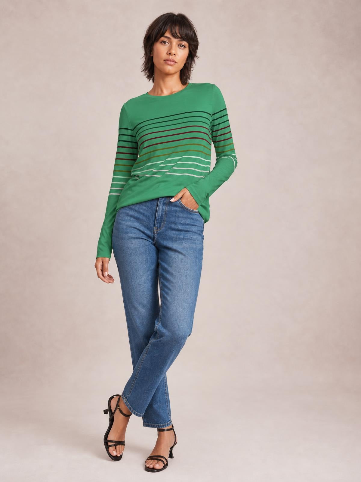 A model poses with one hand in her pocket, wearing a green striped round neck long sleeve T-shirt with a relaxed silhouette and curved horizontal stripes.