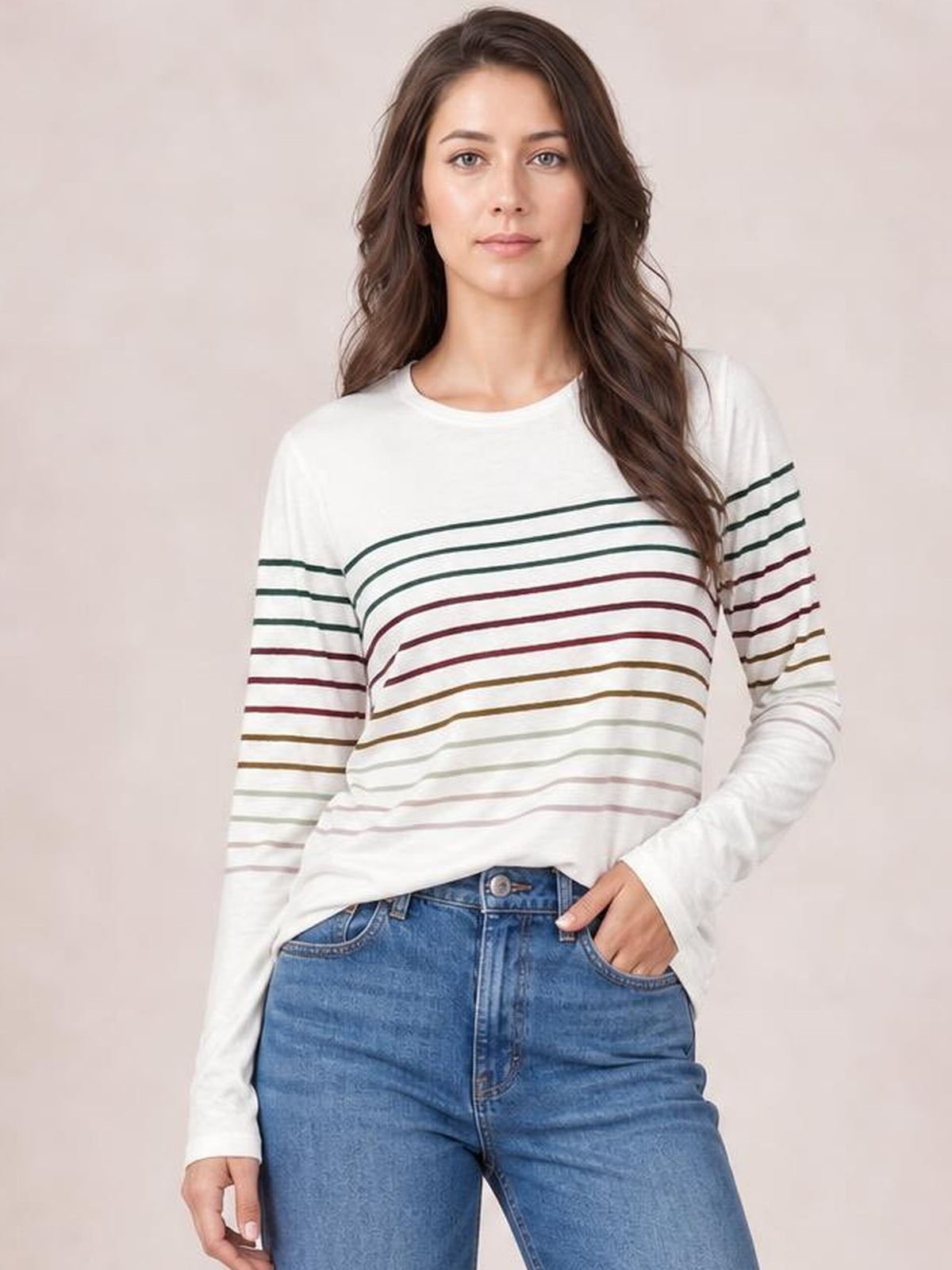 Model wearing a white striped round neck long sleeve T-shirt with multicolor horizontal bands and a relaxed silhouette, posing with one hand in her jeans pocket.