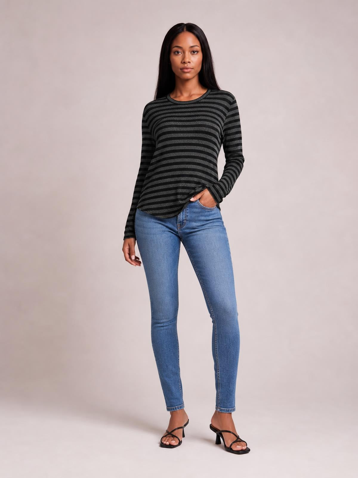 A model stands with one hand in her jeans pocket wearing a black and charcoal striped round-neck long-sleeve T-shirt with a relaxed silhouette and slit hem.