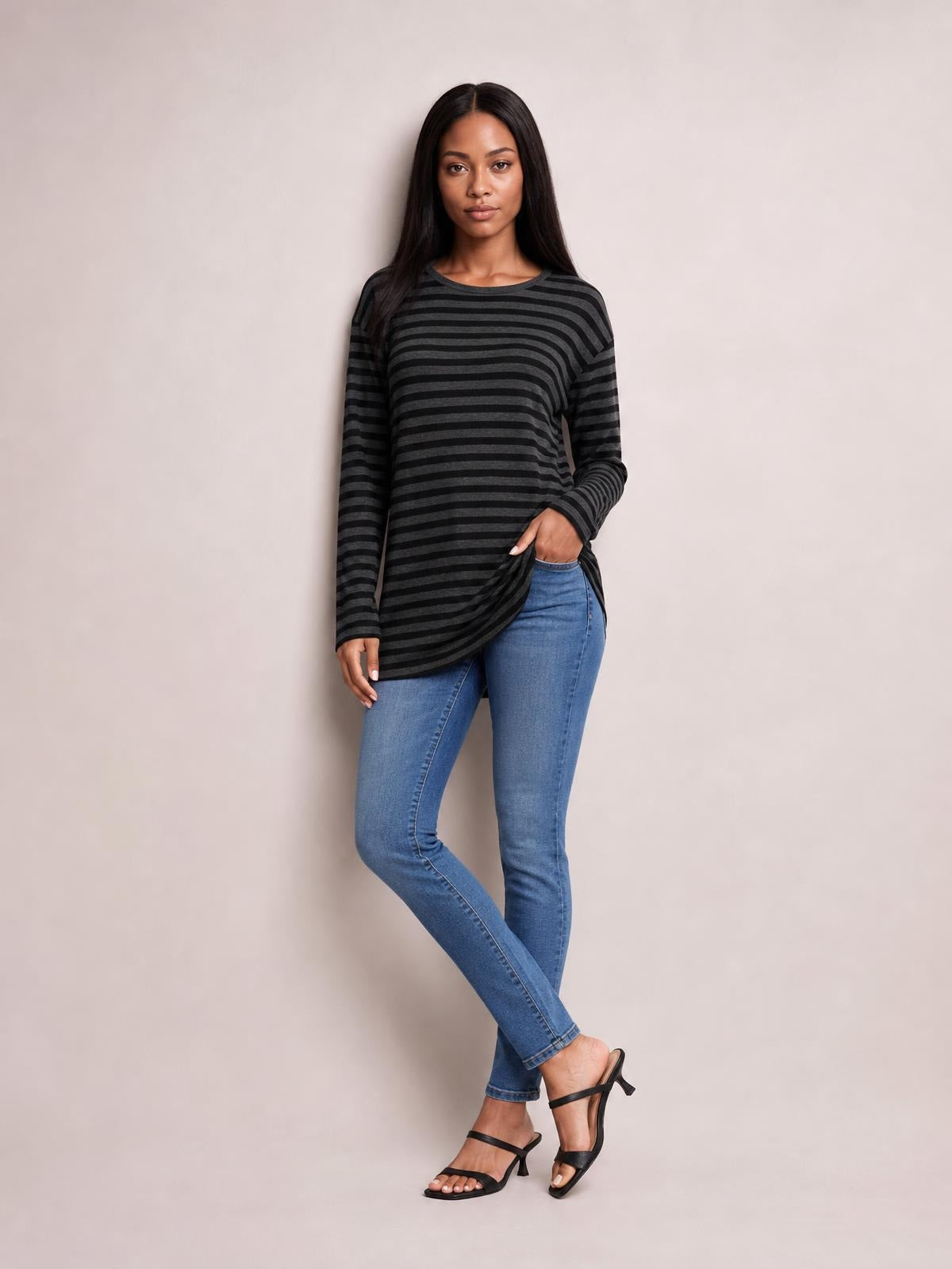 Model poses in a black and charcoal striped round neck long sleeve T-shirt with a relaxed silhouette and side slit detail, paired with jeans.