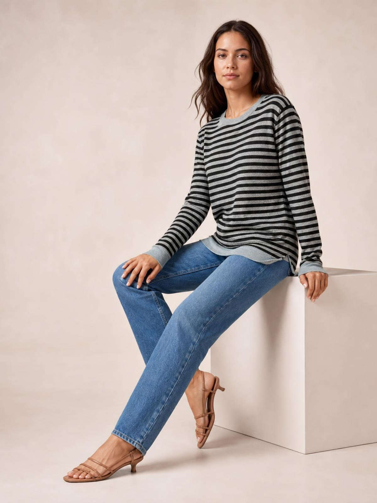 Model seated in a gray and black striped round neck long sleeve T-shirt with a relaxed silhouette, ribbed cuffs and hem, and a visible side slit detail.