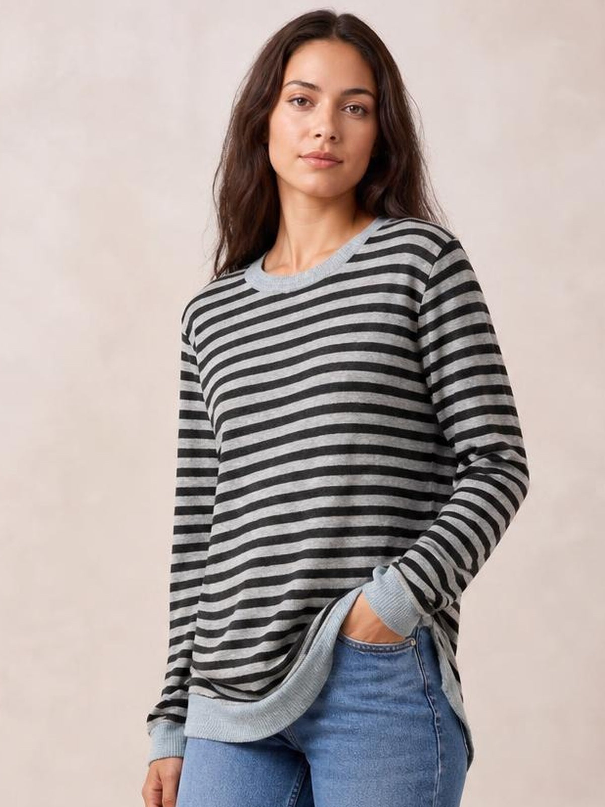 Model wears a loose gray and black striped round neck long sleeve T-shirt with ribbed cuffs and a visible side slit, posing with one hand in her jeans pocket.