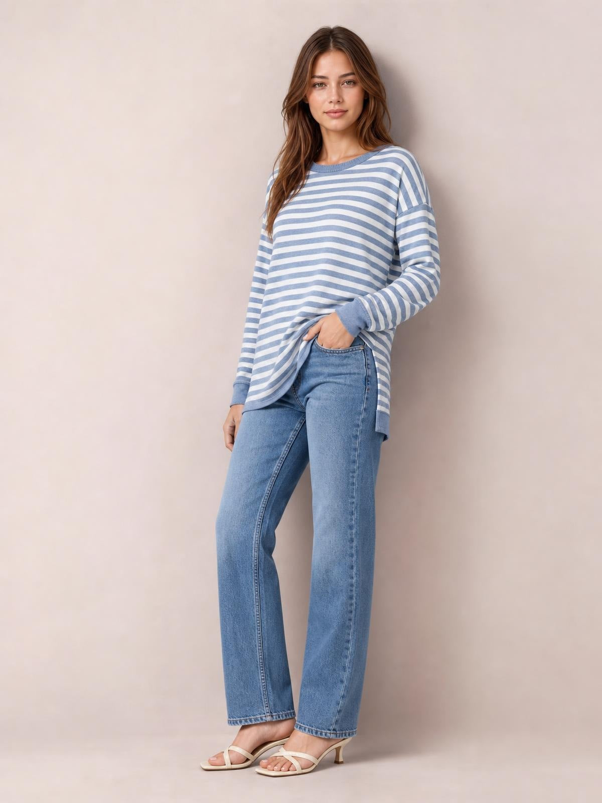 Model wears a blue-and-white striped round neck long sleeve T-shirt with a relaxed silhouette and visible side slit detail, lifting the hem with one hand.
