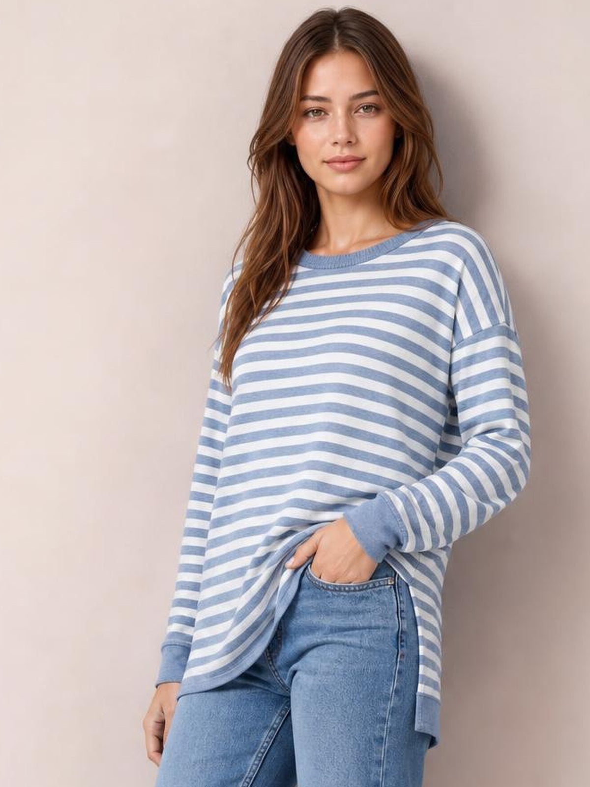 A model poses in a blue and white striped round neck long sleeve T-shirt with a relaxed silhouette, side slit hem, and ribbed cuffs over jeans.