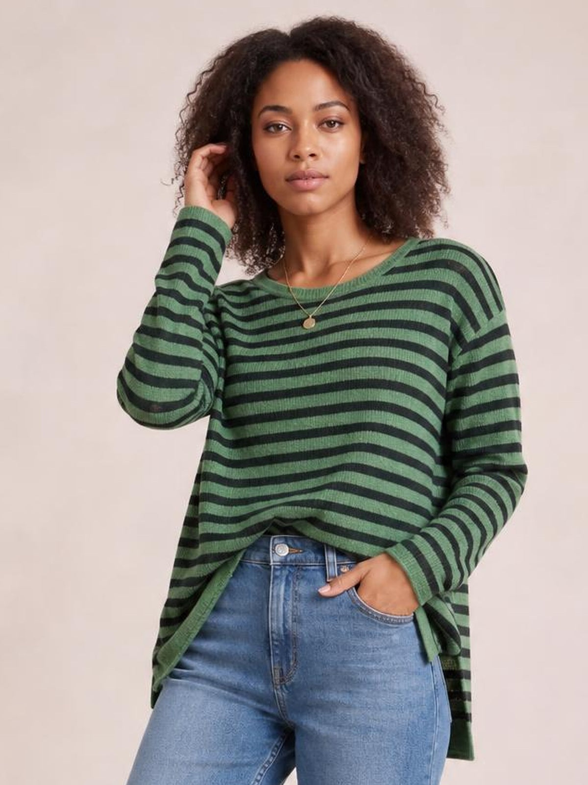 Model wears a green and black striped round-neck long-sleeve T-shirt with a relaxed silhouette, side slit hem, and one hand in her jeans pocket.