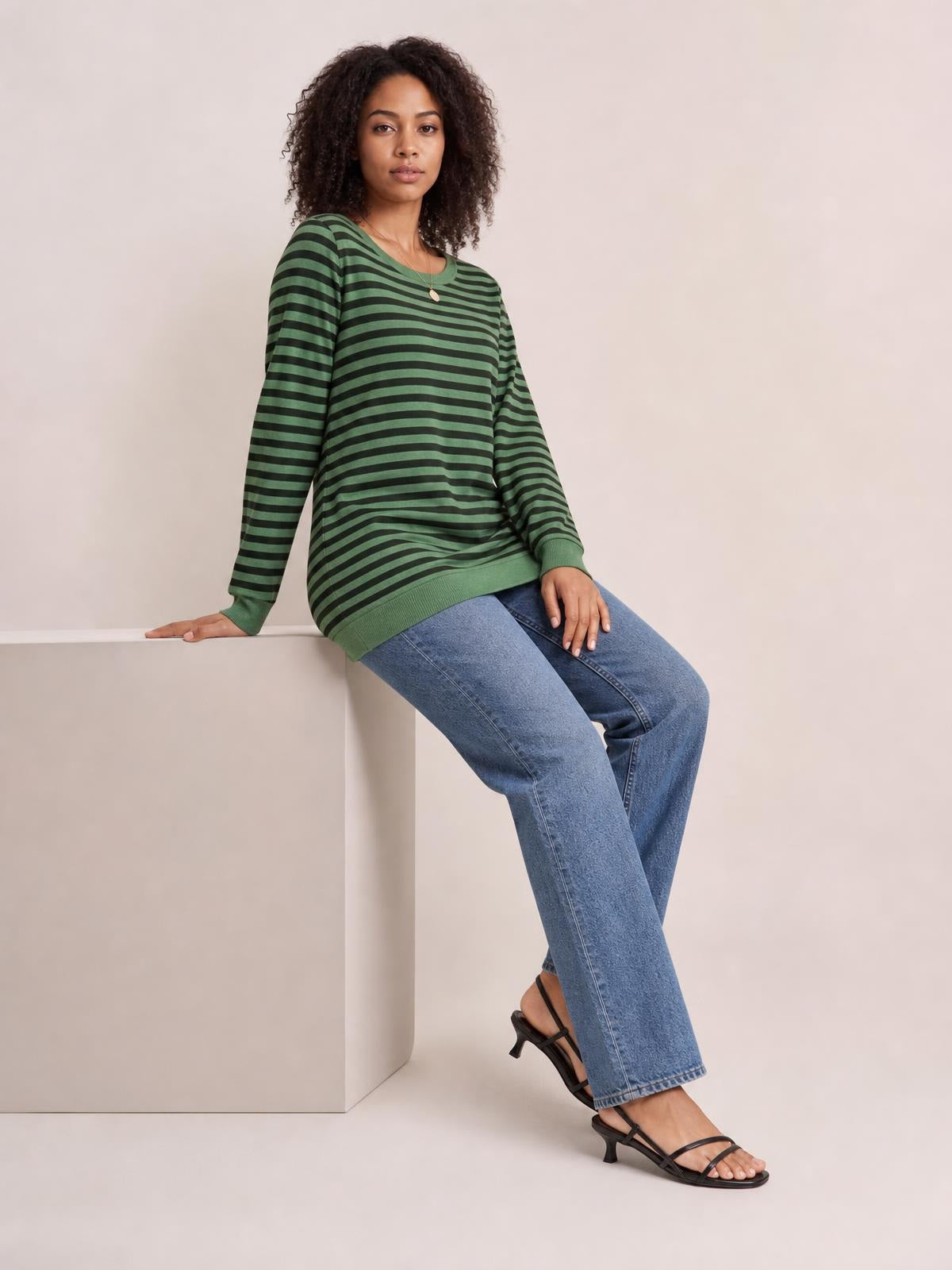 Model seated in a relaxed pose wears a green and black striped round-neck long-sleeve T-shirt with a loose silhouette and ribbed cuffs and hem.