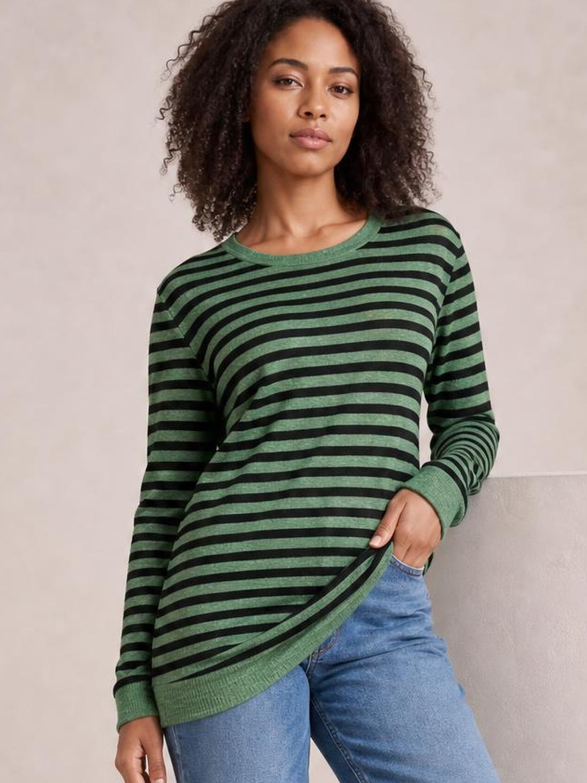 Model in a relaxed pose wears a green and black striped round neck long sleeve T-shirt with a loose silhouette, ribbed cuffs, and a visible side slit.