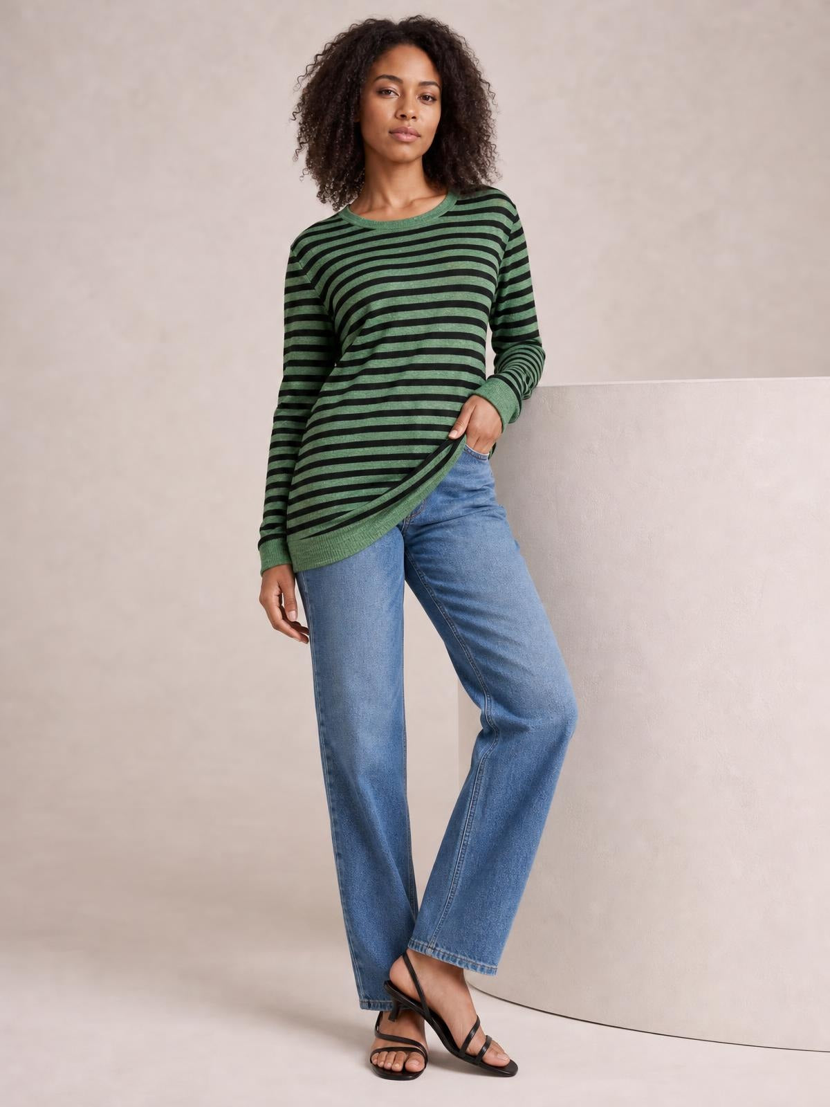 Model wears a green and black striped round neck long sleeve T-shirt with a relaxed silhouette, ribbed trim, and a visible side slit while posing in jeans.