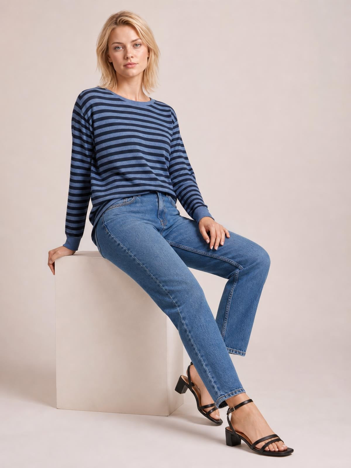 A seated model wears a blue and navy striped round neck long sleeve T-shirt with a relaxed silhouette and visible side slit hem.