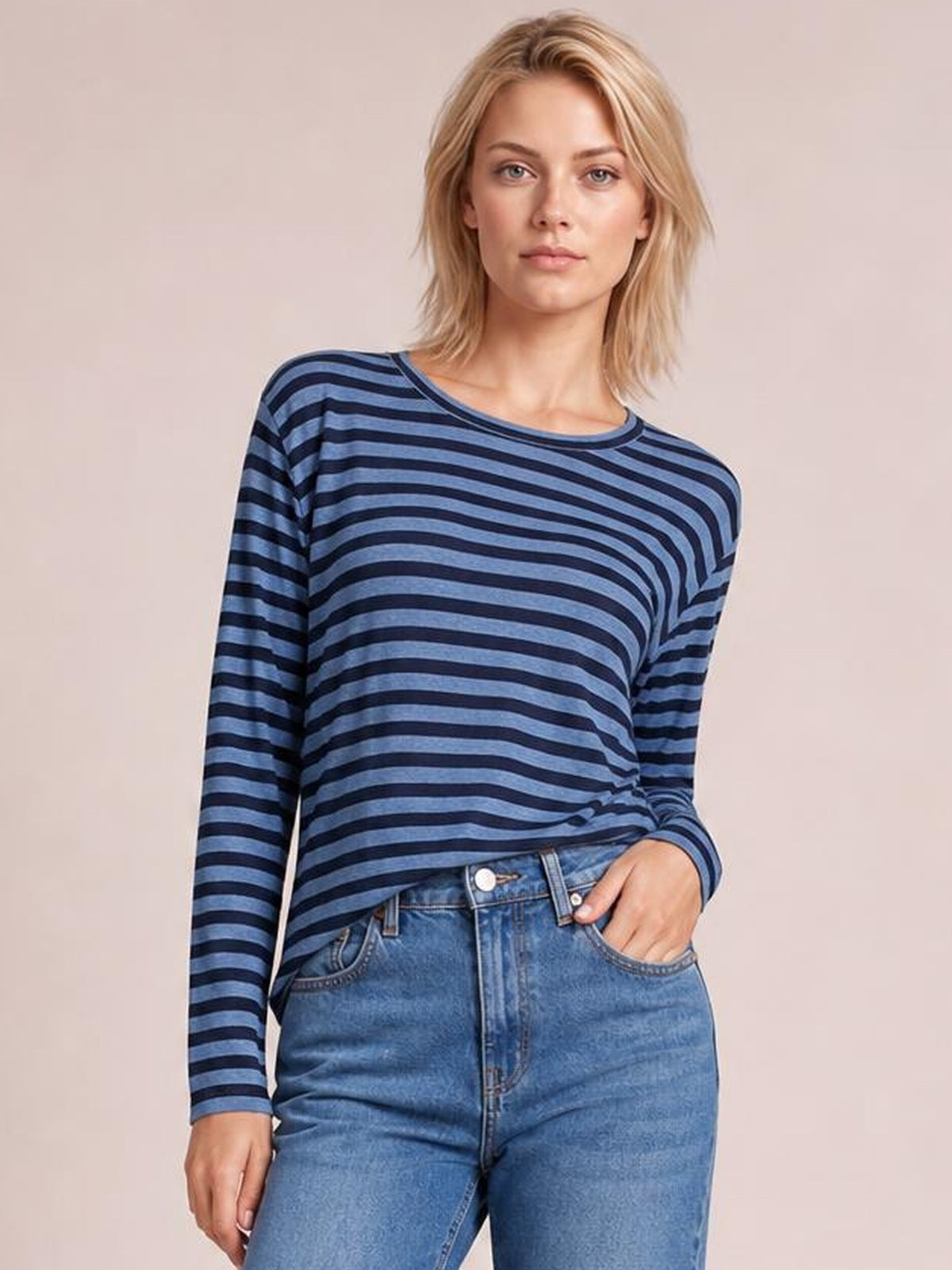 Model wearing a blue and navy striped round neck long sleeve T-shirt with a relaxed silhouette and visible side slit detail, one hand in jeans pocket.