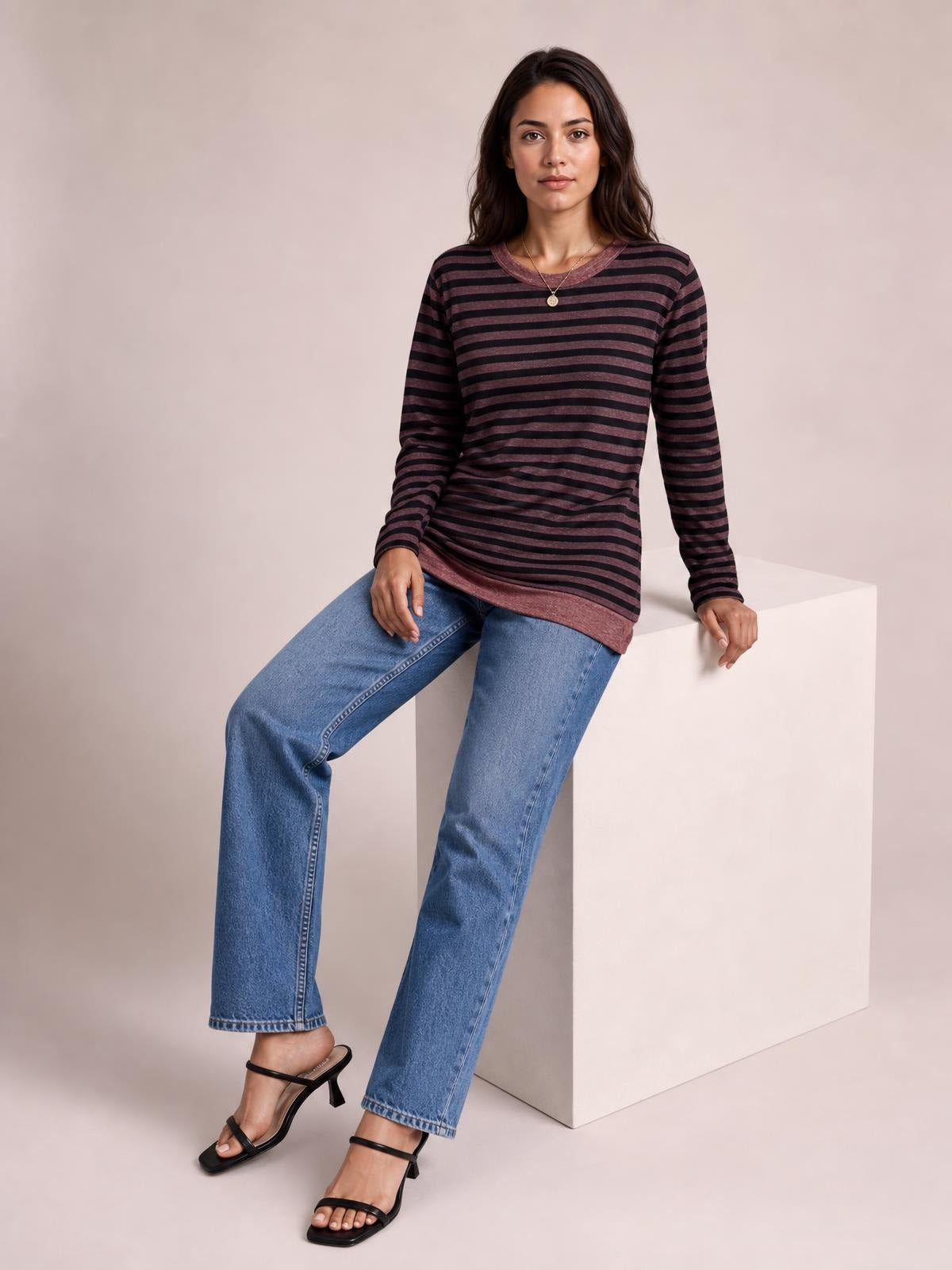 A model sits with one leg extended, wearing a dark plum and black striped round-neck long-sleeve T-shirt with a relaxed silhouette and slit hem detail.